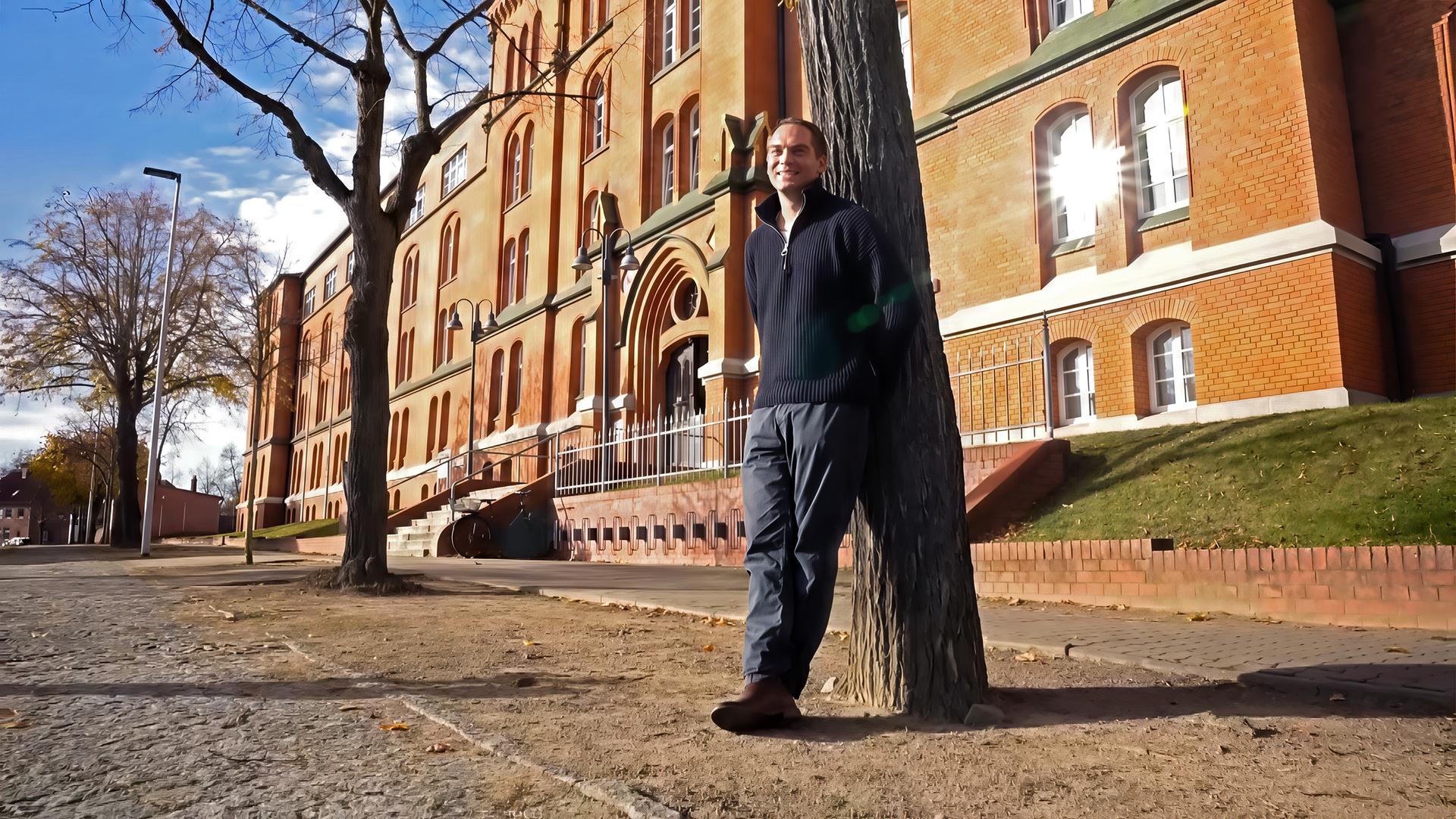 Ein Mann lehnt an einem Baum vor einem alten Backsteingebäude.