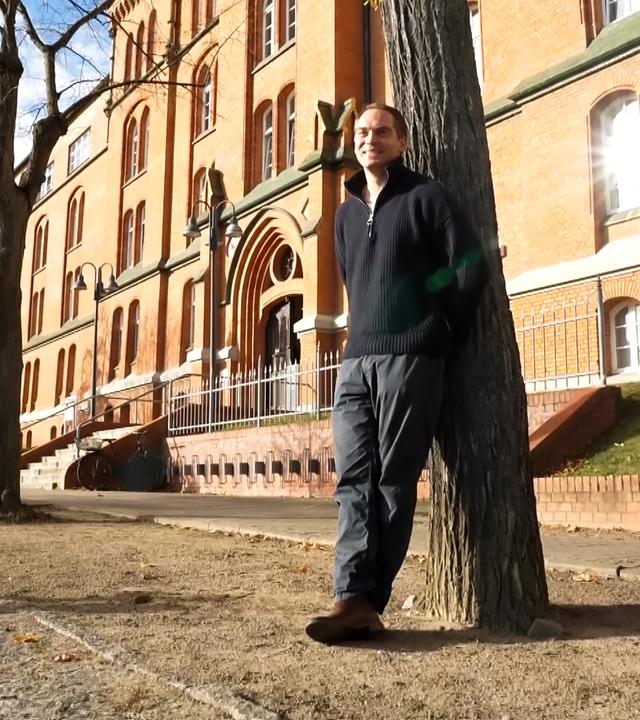 Ein Mann lehnt an einem Baum vor einem alten Backsteingebäude.