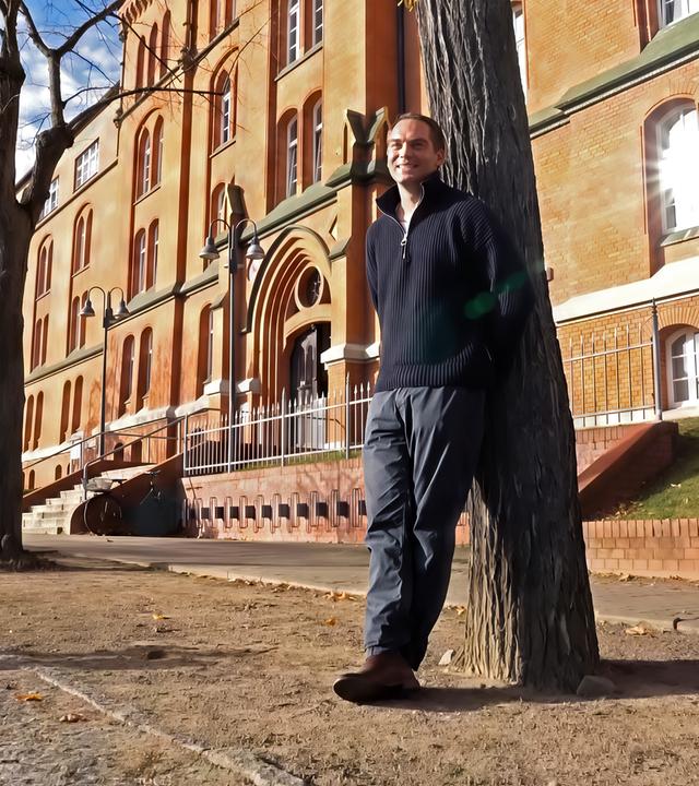 Ein Mann lehnt an einem Baum vor einem alten Backsteingebäude.