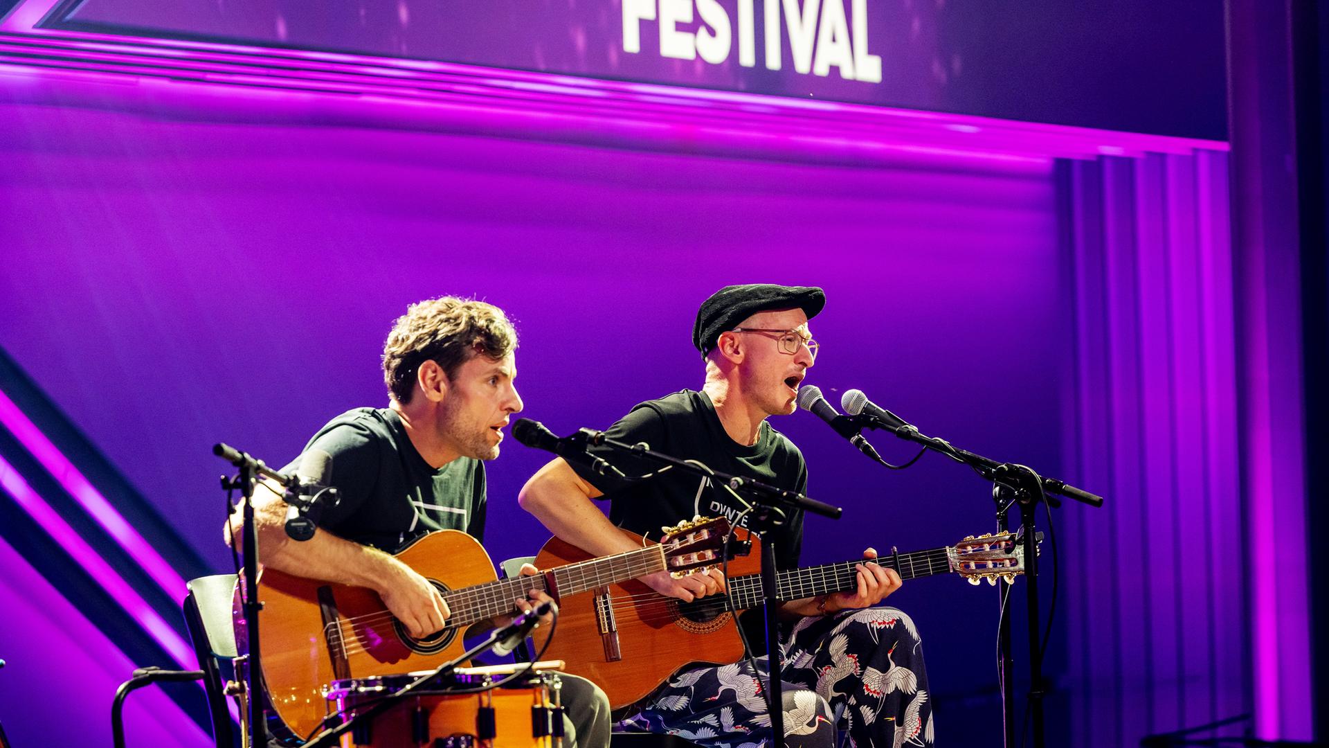 Simon (links) und Jan (rechts) sind auf der Bühne zu sehen. Sie sitzen auf Stühlen in der Mitte des Bildes. Der Hintergrund ist lila beleuchtet.  