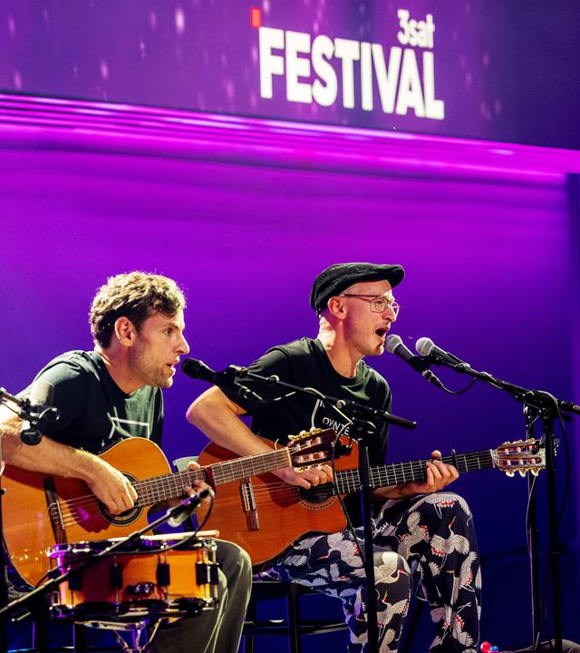 Simon (links) und Jan (rechts) sind auf der Bühne zu sehen. Sie sitzen auf Stühlen in der Mitte des Bildes. Der Hintergrund ist lila beleuchtet.  