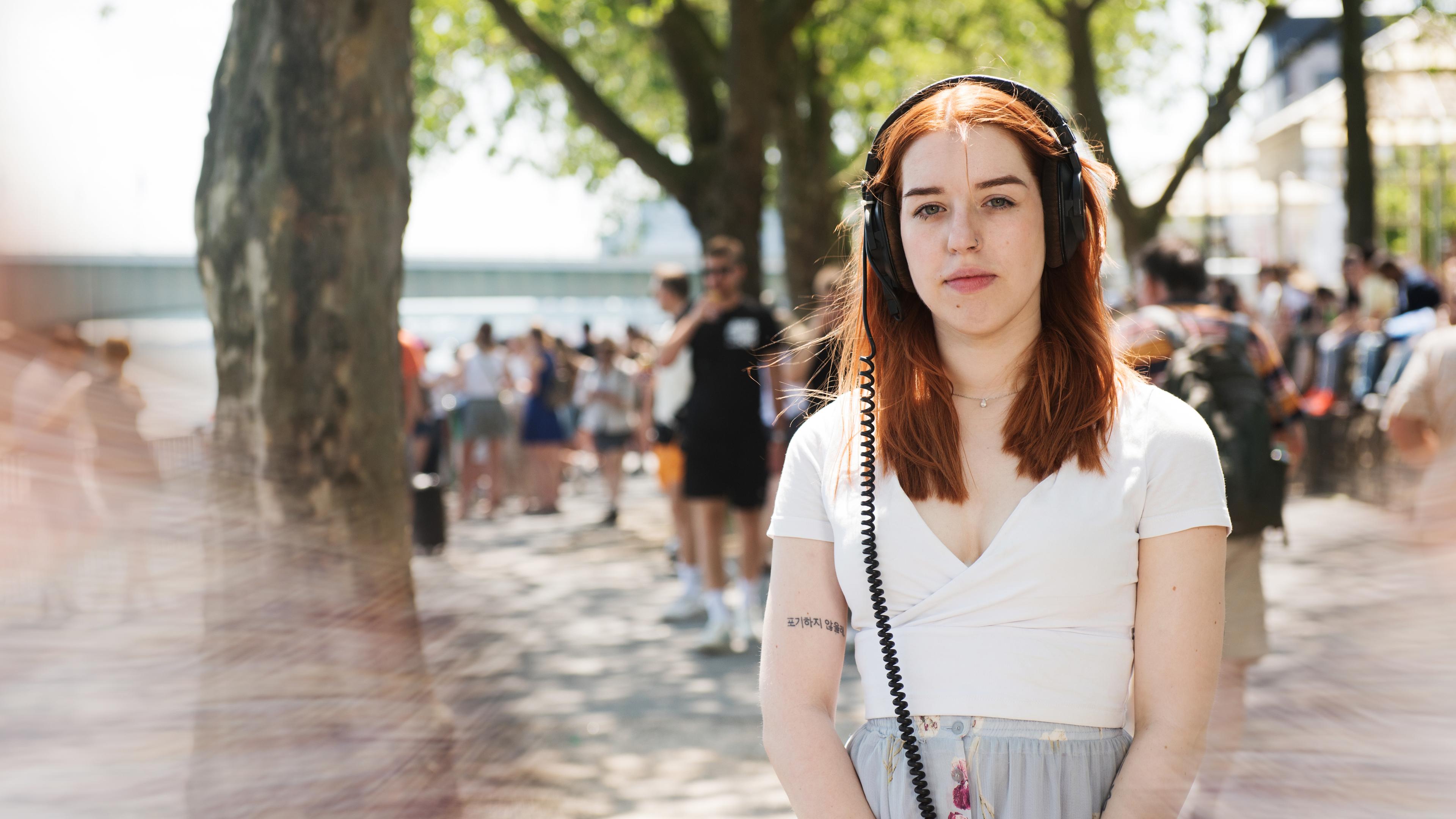 Jungefrau mit langen rotbraunen, glatten Haaren und Kopfhörern auf den Ohren steht in einer Fußgängerzone. Hintergrund unscharf. Sie schaut ernst in die Kamera.