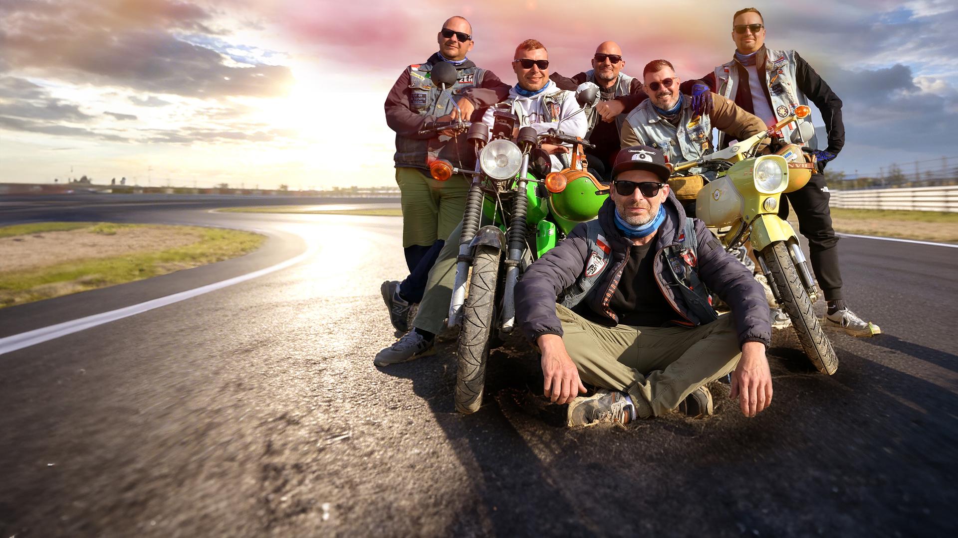 Eine Gruppe von sieben Männern mit zwei Simson-Mopeds auf einer Landstraße. Die Männer tragen Sonnenbrille und lächeln in die Kamera, einer sitzt vor der stehenden Gruppe im Schneidersitz auf der Straße.