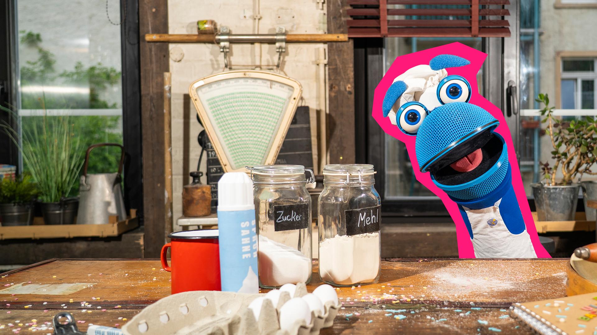 SingDing schaut hinter einem Tisch voller Backutensilien hervor. Vor ihm stehen eine offene Eierpackung, Sahne, Zucker und Mehl, ein Backbuch und ein Nudelholz.