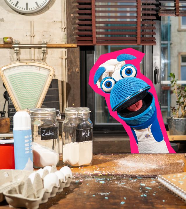 SingDing schaut hinter einem Tisch voller Backutensilien hervor. Vor ihm stehen eine offene Eierpackung, Sahne, Zucker und Mehl, ein Backbuch und ein Nudelholz.