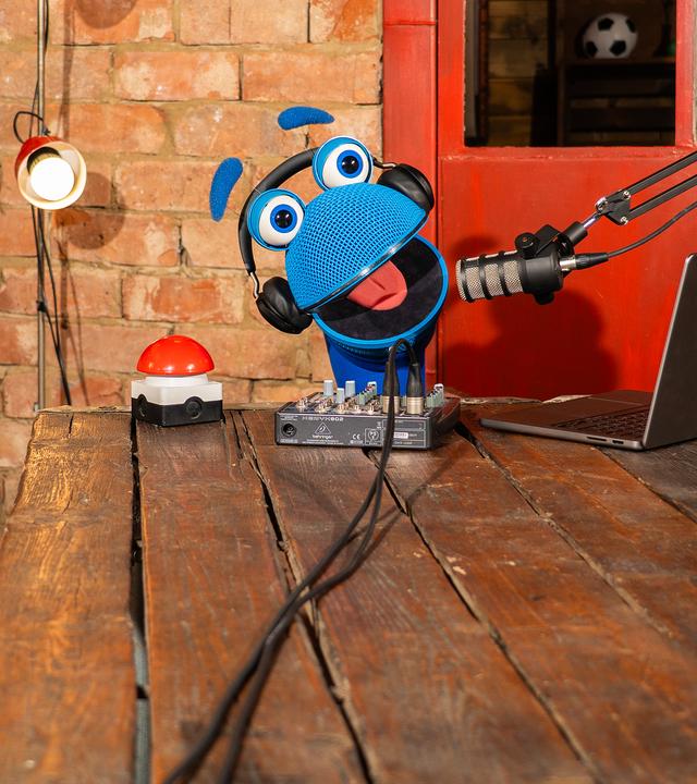 "SingAlarm - Radio SingAlarm": SingDing schaut hinter einem Holztisch hervor. Es trägt Kopfhörer und spricht in ein Standmikrofon. Vor ihm auf dem Tisch stehen ein roter Buzzer, ein Tonmischer und ein Laptop.