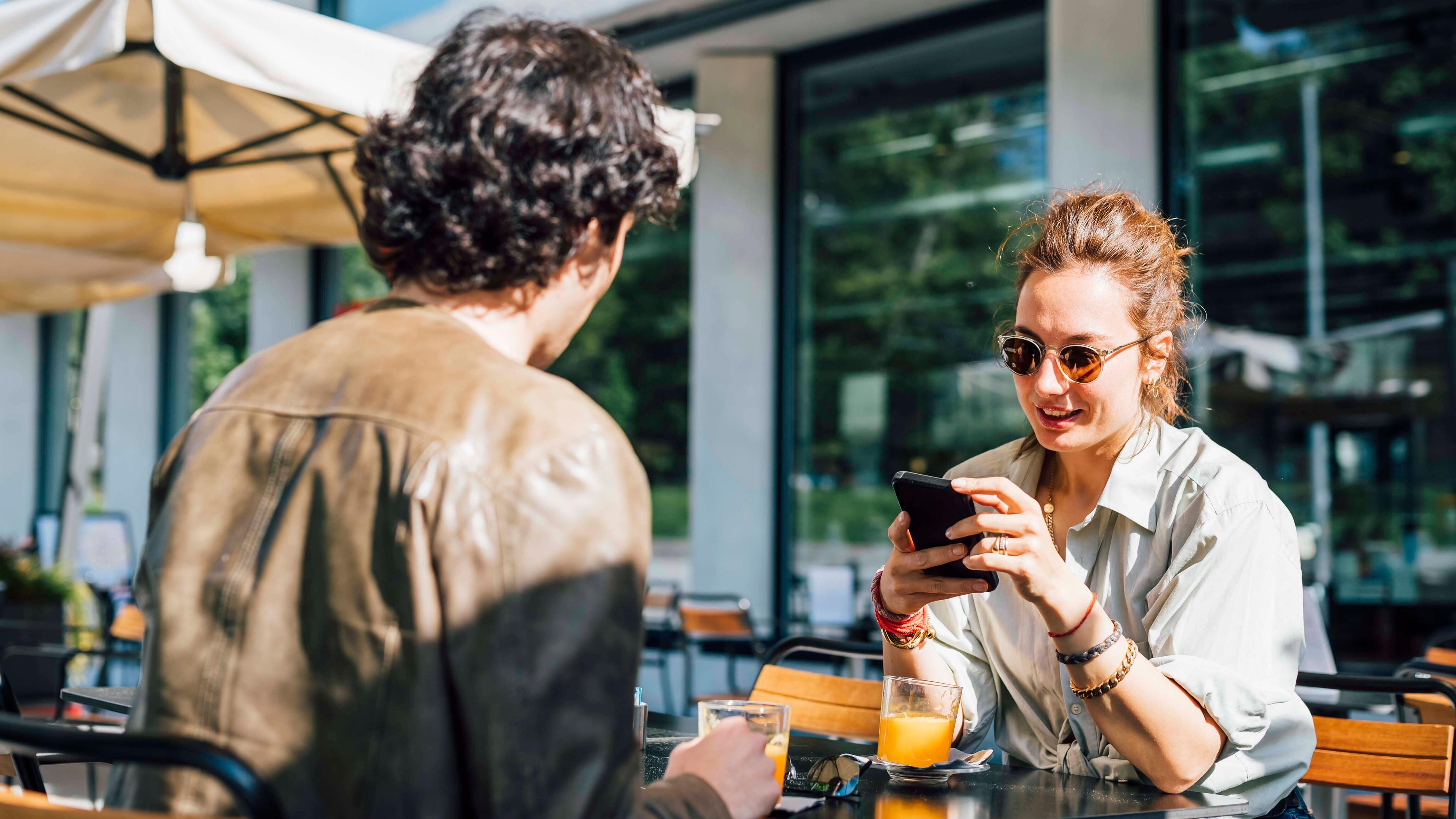 Ein Pärchen, bestehend aus Frau und Mann, sitzt im Außenbereich eines Cafés. Die Frau hält ein Smartphone in der Hand.