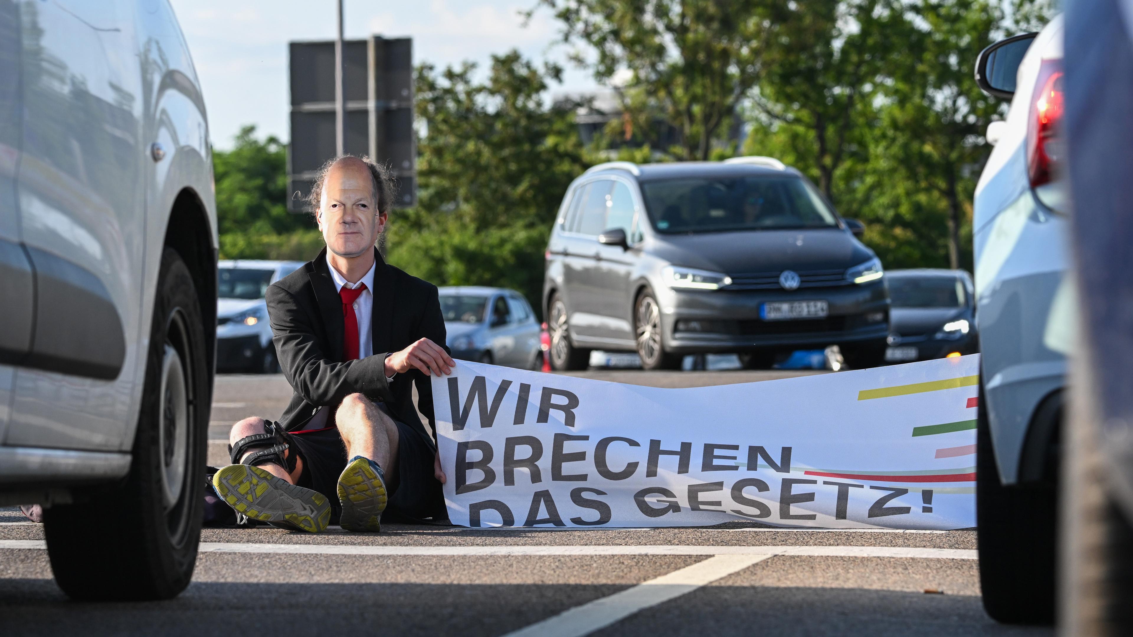 Ein Mitglied der Protestgruppe Letzte Generation nimmt an einer Sitzblockade an der Autobahnabfahrt am Elbepark teil und trägt dabei eine Maske mit dem Porträt von Bundeskanzler Scholz.
