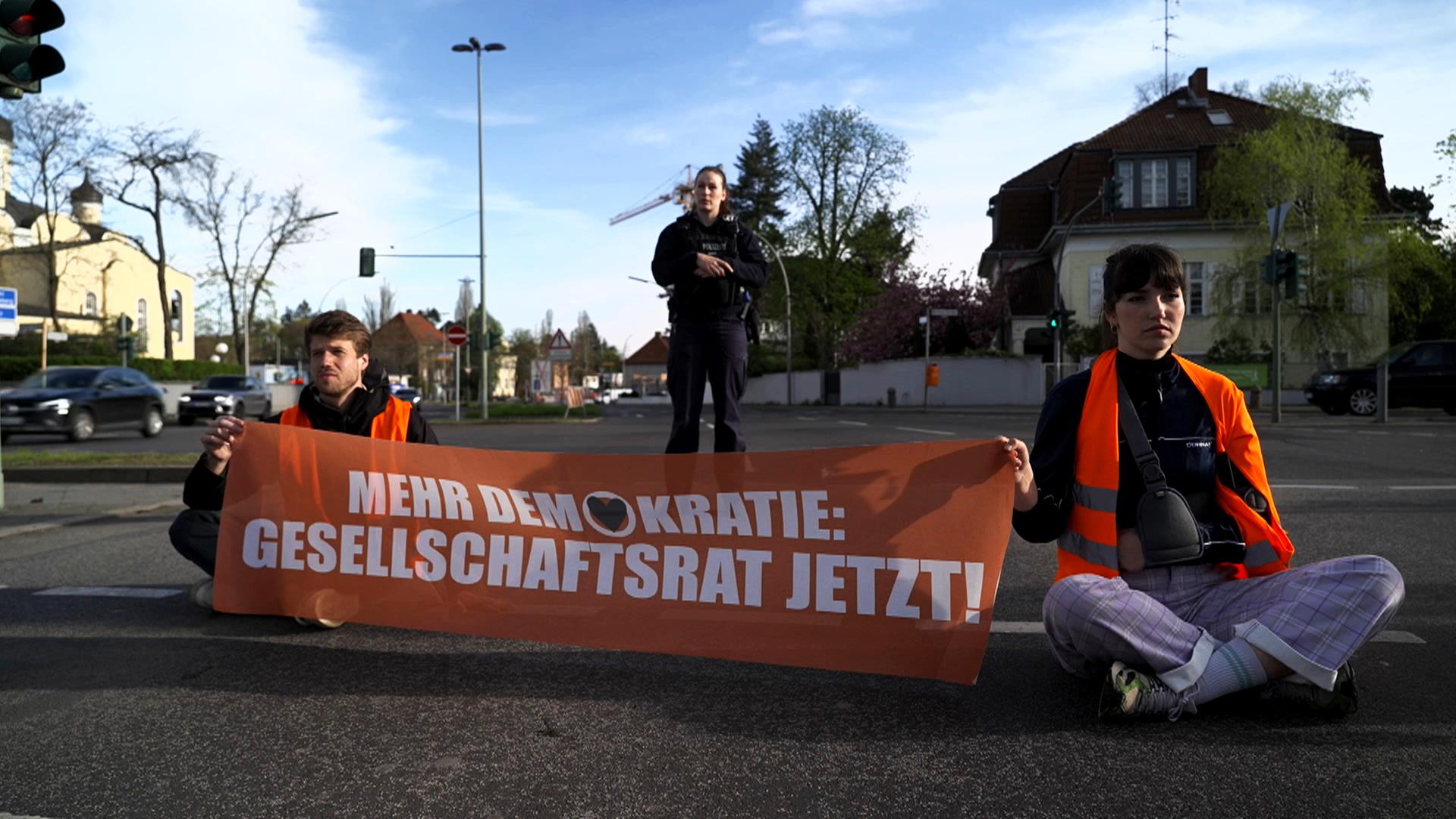 Sitzblockade Klimakleber der "Letzten Generation" an Verkehrskreuzung mit Transparent "Mehr Demokratie: Gesellschaftsrat jetzt!"