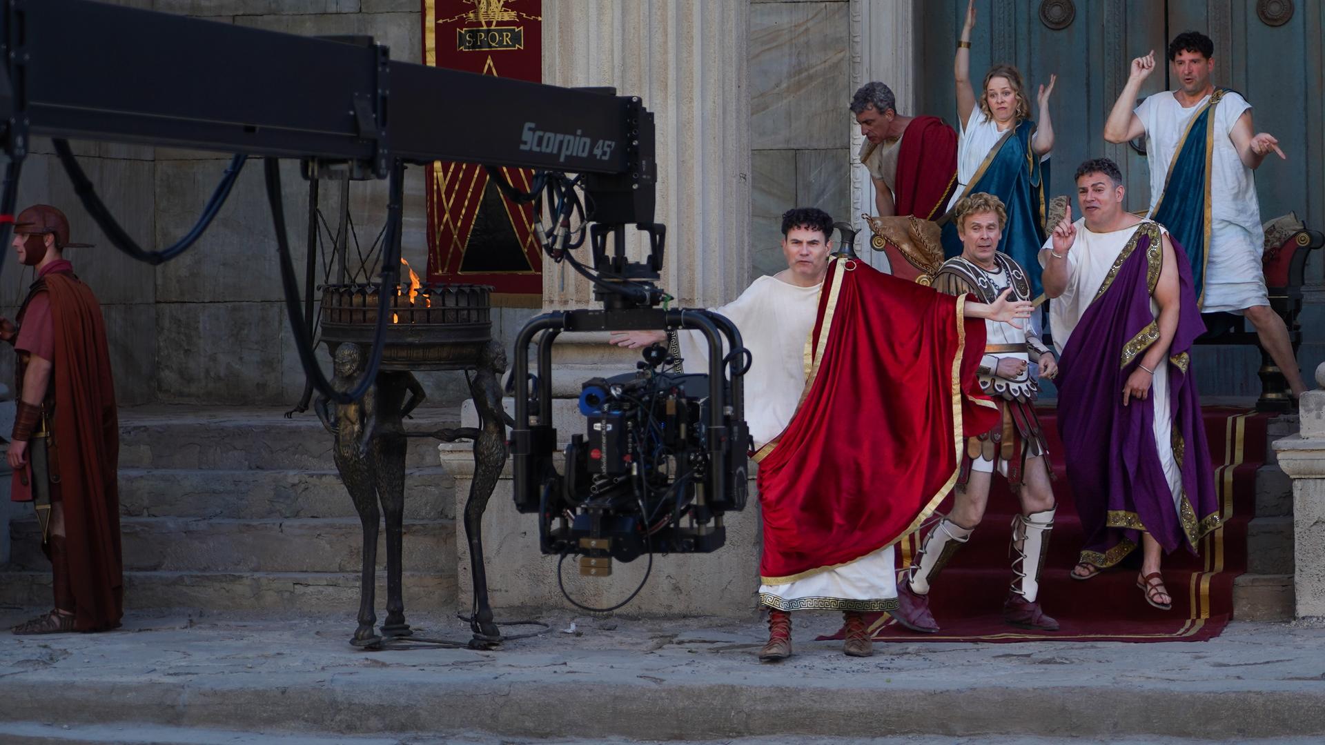f den Treppen vor einem Eingangsportal zu einem großen Gebäude stehen Alexander Schubert, Paul Sedlmeir, Jasmin Schwiers, Matthias Matschke und Carsten Strauch. Sie alle haben beim Tanzen unterschiedliche Posen eingenommen. Vor ihnen ragt ein Kameraarm ins Bild, neben ihnen stehen Feuerschalen.