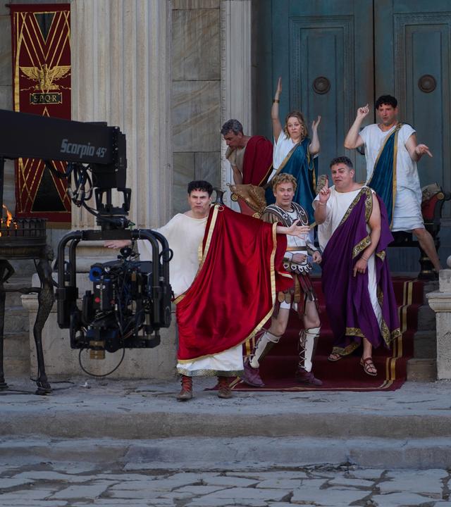 f den Treppen vor einem Eingangsportal zu einem großen Gebäude stehen Alexander Schubert, Paul Sedlmeir, Jasmin Schwiers, Matthias Matschke und Carsten Strauch. Sie alle haben beim Tanzen unterschiedliche Posen eingenommen. Vor ihnen ragt ein Kameraarm ins Bild, neben ihnen stehen Feuerschalen.