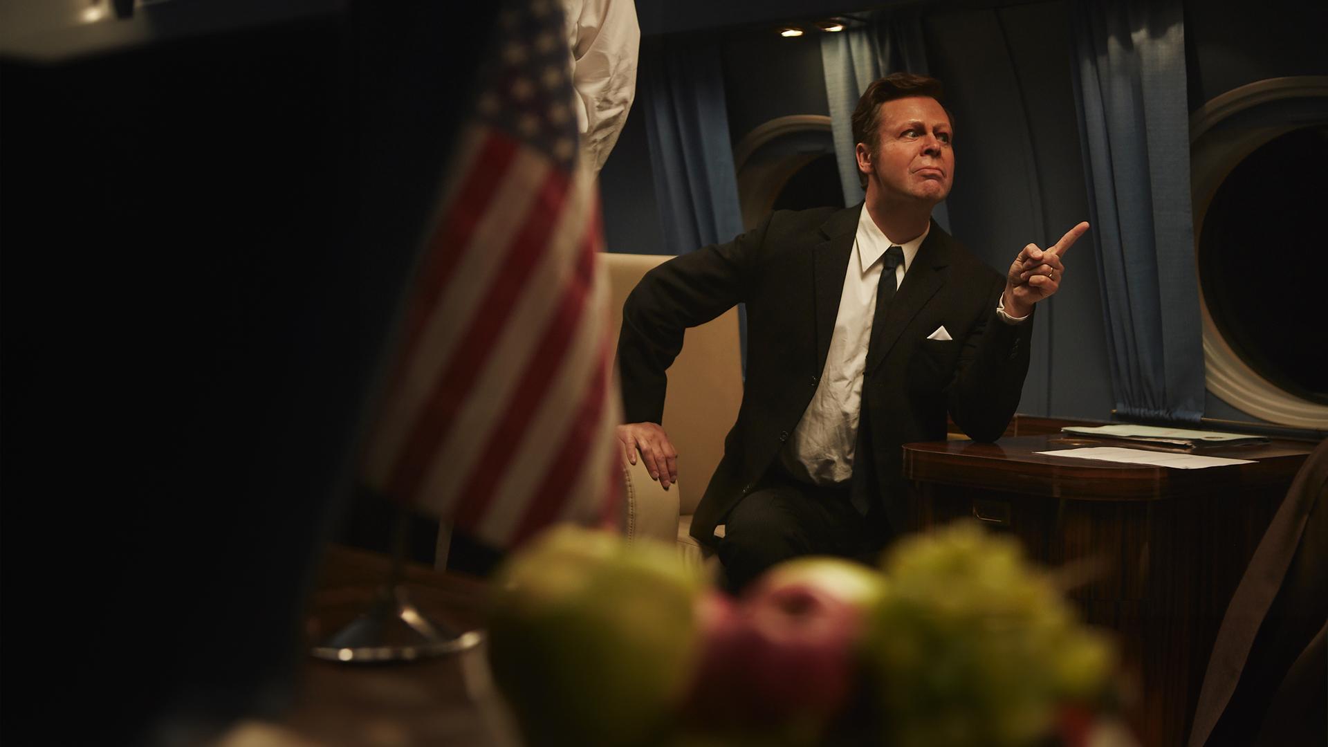 Sketch History USA Spezial: Matthias Matschke sitzt als John F. Kennedy maskiert auf einem Flugzeugsitz, streckt seinen Zeigefinger seinem Gegenüber entgegen.