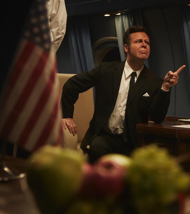 Sketch History USA Spezial: Matthias Matschke sitzt als John F. Kennedy maskiert auf einem Flugzeugsitz, streckt seinen Zeigefinger seinem Gegenüber entgegen.