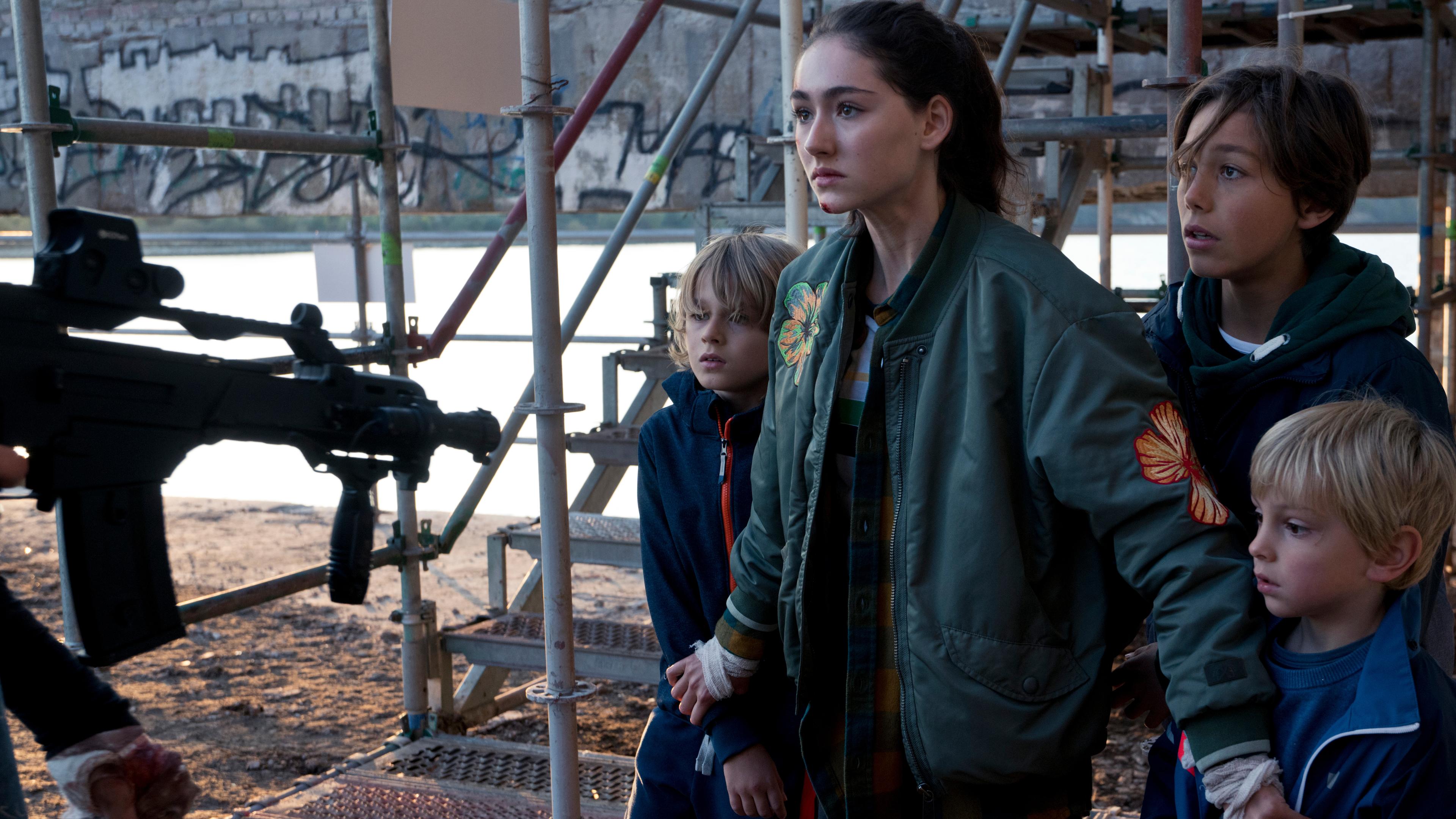 "Sløborn": Evelin (Emily Kusche) steht schützend vor ihren Brüdern (Maximilian Brauer, Phileas Heyblom, Ron Renzenbrink). Am Bildrand ist zu sehen, dass sie von einem Maschinengewehr bedroht werden.