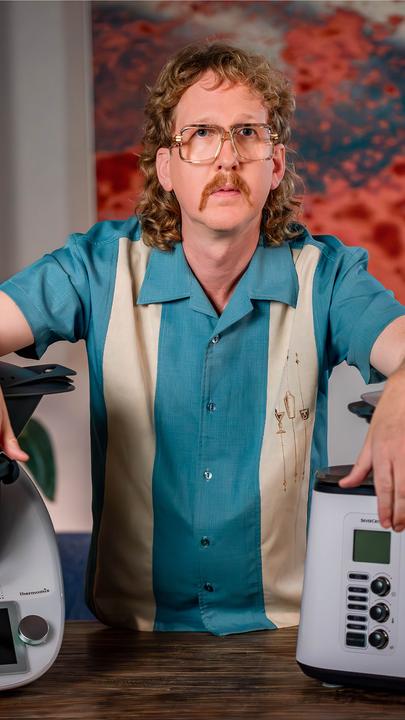 Uke Bosse schaut ernst in die Kamera, seine Arme stützen sich links auf einen Thermomix und rechts auf eine "Monsieur Cuisine"-Maschine.