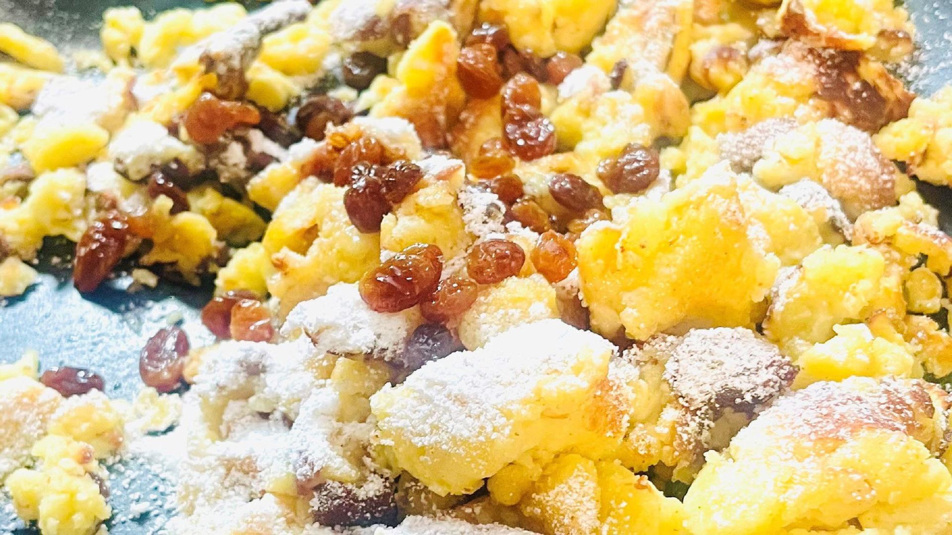 Das Bild zeigt eine eine Portion Kaiserschmarrn, einem traditionellen österreichischen Gericht. Der Kaiserschmarrn besteht aus grob zerrissenen, goldbraun gebackenen Teigstücken, die mit Puderzucker bestäubt sind. Darüber befinden sich einige Rosinen, die in den Teig eingearbeitet sind. Die Stücke sind unregelmäßig geformt und leicht angebräunt an den Rändern. Der Hintergrund des Bildes ist eine dunkle Pfanne, die den Kaiserschmarrn präsentiert. Der Gesamteindruck ist appetitlich und die Farben variieren von goldgelb bis braun, begleitet von dem weißen Puderzucker.