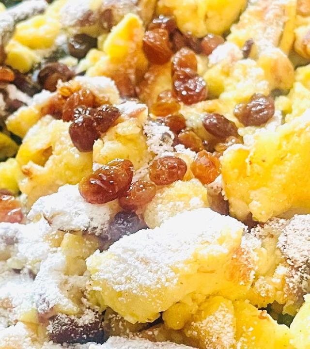 Das Bild zeigt eine eine Portion Kaiserschmarrn, einem traditionellen österreichischen Gericht. Der Kaiserschmarrn besteht aus grob zerrissenen, goldbraun gebackenen Teigstücken, die mit Puderzucker bestäubt sind. Darüber befinden sich einige Rosinen, die in den Teig eingearbeitet sind. Die Stücke sind unregelmäßig geformt und leicht angebräunt an den Rändern. Der Hintergrund des Bildes ist eine dunkle Pfanne, die den Kaiserschmarrn präsentiert. Der Gesamteindruck ist appetitlich und die Farben variieren von goldgelb bis braun, begleitet von dem weißen Puderzucker.