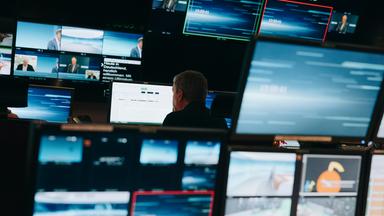 Eine Vielzahl blauer Bildschirme umrahmen einen Mitarbeiter in einer Fernsehregie 