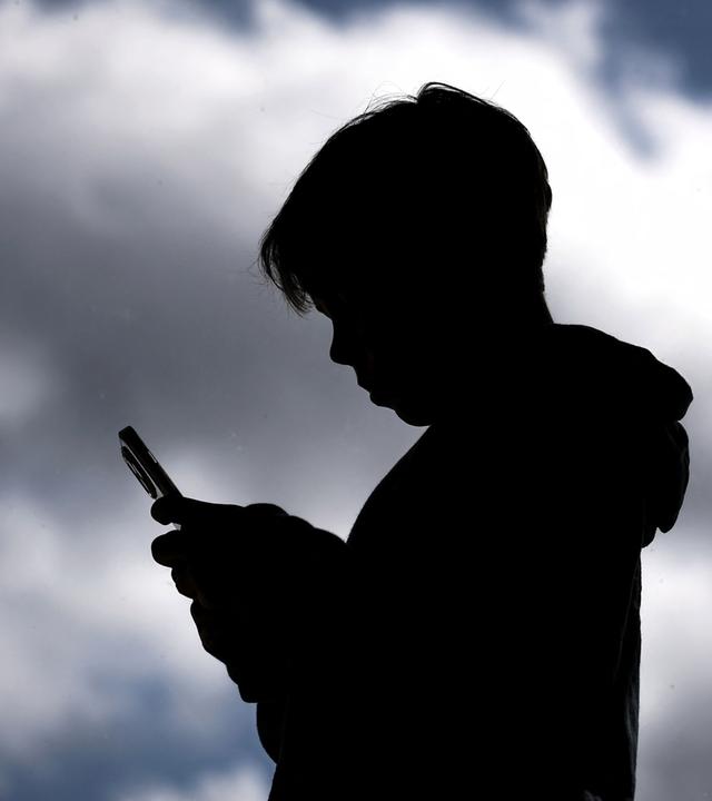 Jugendlicher schaut auf sein Smartphone, Schattenriss vor dunklen Wolken.