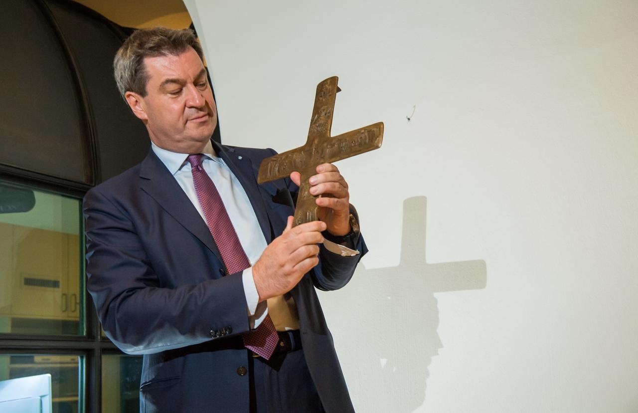 Markus Söder steht in einem Raum und hält ein Kreuz hoch.