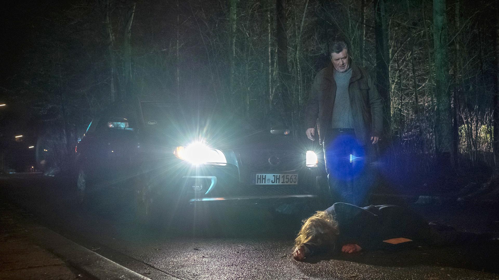 "Notruf Hafenkante - SOKO Kiebitz": Hans Moor (Bruno F. Apitz) steht im Dunkeln auf einer Landstraße vor seinem Auto und schaut entsetzt auf eine Frau (Tanja Schleiff), die auf der Straße vor ihm liegt. Im Hintergrund ist Wald zu sehen.