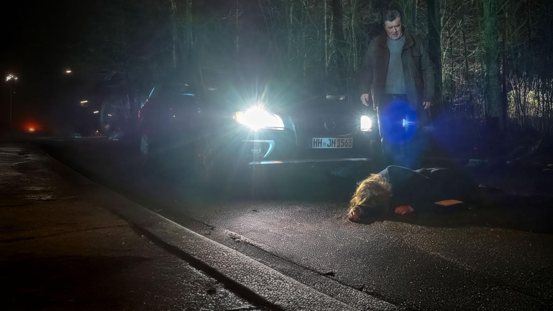 "Notruf Hafenkante - SOKO Kiebitz": Hans Moor (Bruno F. Apitz) steht im Dunkeln auf einer Landstraße vor seinem Auto und schaut entsetzt auf eine Frau (Tanja Schleiff), die auf der Straße vor ihm liegt. Im Hintergrund ist Wald zu sehen.