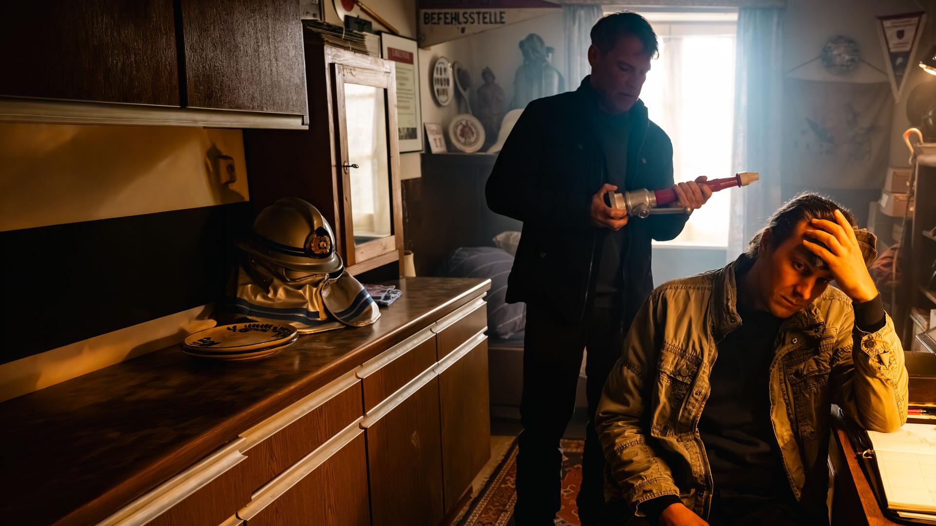 "SOKO Köln - Feuer und Flamme": Nils Steiner (Frederik Götz) sitzt an einem braunen Schreibtisch und hat den Kopf in seine linke Hand gestützt. Er sitzt in Richtung des Betrachters und blickt traurig nach unten. Hinter ihm steht Matti Wagner (Pierre Besson) und hält einen länglichen Gegenstand in den Händen, den er interessiert betrachtet. Eine Lampe über dem Schreibtisch spendet warmes Licht. Durch ein Fenster hinter Matti fällt Tageslicht in den Raum.