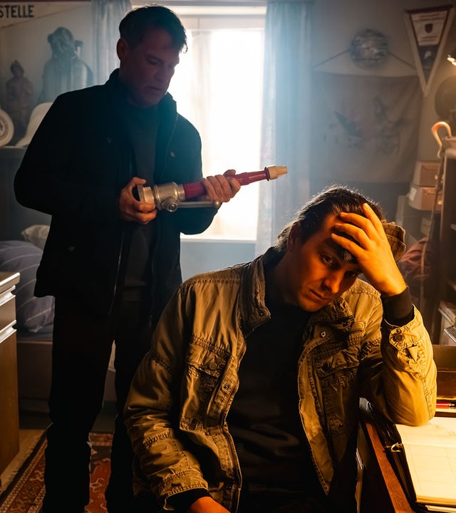 "SOKO Köln - Feuer und Flamme": Nils Steiner (Frederik Götz) sitzt an einem braunen Schreibtisch und hat den Kopf in seine linke Hand gestützt. Er sitzt in Richtung des Betrachters und blickt traurig nach unten. Hinter ihm steht Matti Wagner (Pierre Besson) und hält einen länglichen Gegenstand in den Händen, den er interessiert betrachtet. Eine Lampe über dem Schreibtisch spendet warmes Licht. Durch ein Fenster hinter Matti fällt Tageslicht in den Raum.