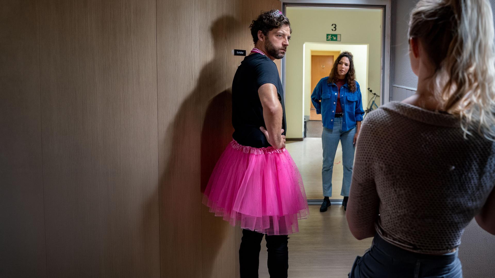 "SOKO Leipzig - Auf Sand gebaut": Johannes Schäfer (Timur Isik) steht in einem Türrahmen und schaut zu seiner Frau Judith (Anne Kanis), die nur im Anschnitt von hinten zu sehen ist. Er trägt ein rosa Tutu und auf dem Kopf ein Spielzeugdiadem. Kim Nowak (Amy Mußul) steht neben Johannes und sieht ihn überrascht an.