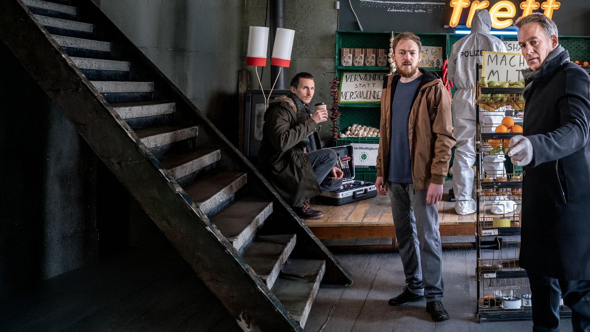 "SOKO Leipzig - Rette sich, wer kann": Moritz Brenner (Johannes Hendrik Langer) hockt mit einem Coffee-to-go-Becher im Hintergrund des Ladengeschäfts und sieht zu Simon Koschanowsky (Tino Mewes) und Jan Maybach (Marco Girnth). Die beiden stehen vor ihm im Geschäft. Simon schaut in die Richtung, in die Jan deutet.