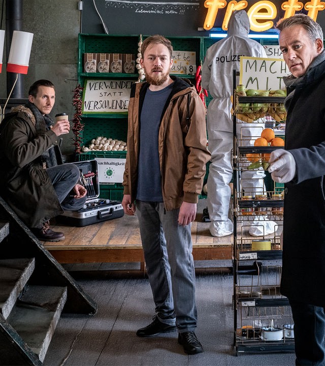 "SOKO Leipzig - Rette sich, wer kann": Moritz Brenner (Johannes Hendrik Langer) hockt mit einem Coffee-to-go-Becher im Hintergrund des Ladengeschäfts und sieht zu Simon Koschanowsky (Tino Mewes) und Jan Maybach (Marco Girnth). Die beiden stehen vor ihm im Geschäft. Simon schaut in die Richtung, in die Jan deutet.