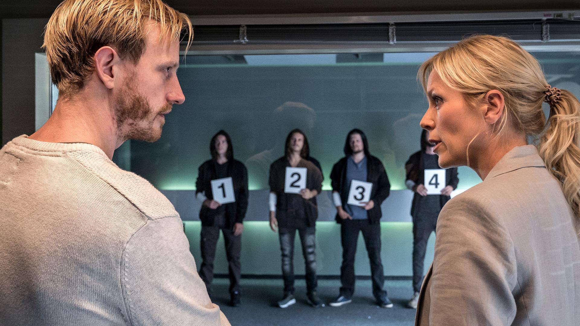 Peter Hassmann (David Zimmerschied) steht mit verschränkten Armen neben Ina Zimmermann (Melanie Marschke) vor einer Glasscheibe