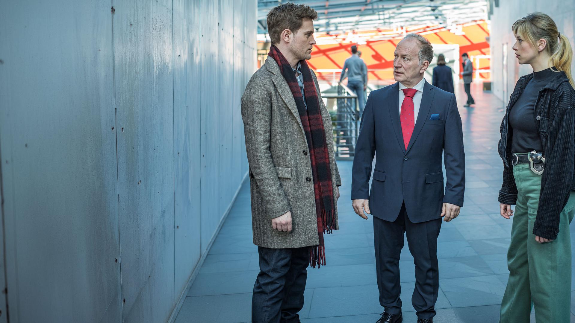 "SOKO München - Affenliebe": Die Kommissare Resi Schwaiger (Mersiha Husagic) und Dominik Morgenstern (Joscha Kiefer) stehen mit Gerhard Beck (Stefan Merki), dem Geschäftsführer des Institutes, an dem das Opfer studiert hat, im Flur. Beck trägt einen edlen Anzug und steht zwischen den beiden Kommissaren, während er Dominik Morgenstern entsetzt in die Augen schaut. Resi Schwaiger sieht währenddessen zu ihrem Kollegen.