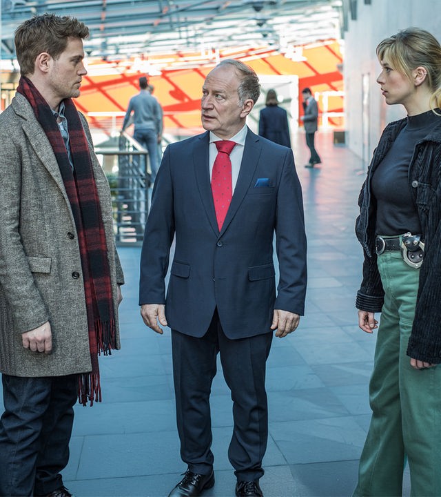 "SOKO München - Affenliebe": Die Kommissare Resi Schwaiger (Mersiha Husagic) und Dominik Morgenstern (Joscha Kiefer) stehen mit Gerhard Beck (Stefan Merki), dem Geschäftsführer des Institutes, an dem das Opfer studiert hat, im Flur. Beck trägt einen edlen Anzug und steht zwischen den beiden Kommissaren, während er Dominik Morgenstern entsetzt in die Augen schaut. Resi Schwaiger sieht währenddessen zu ihrem Kollegen.