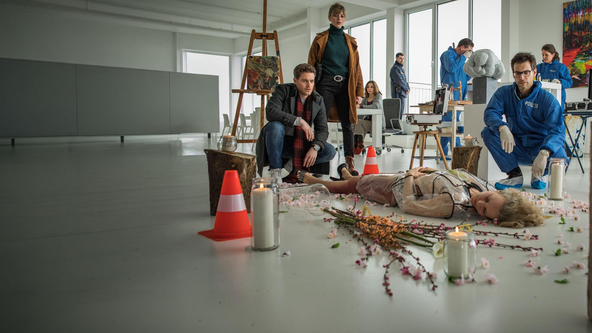 "SOKO München - Ein kunstvoller Tod": In einer Galerie liegt eine weibliche Leiche auf dem Boden um sie herum sind Blumen und Kerzengläser verteilt. Dr. Weissenböck (Florian Odendahl) sitzt in der Hocke neben ihr und schaut sie an. Am Fußende sitzt Dominik Morgenstern (Joscha Kiefer) in der Hocke, neben ihm steht Resi Schwaiger (Mersiha Husagic), sie schauen ebenfalls auf die Leiche. Im Hintergrund ist die Spusi (Komparsen) zu sehen.