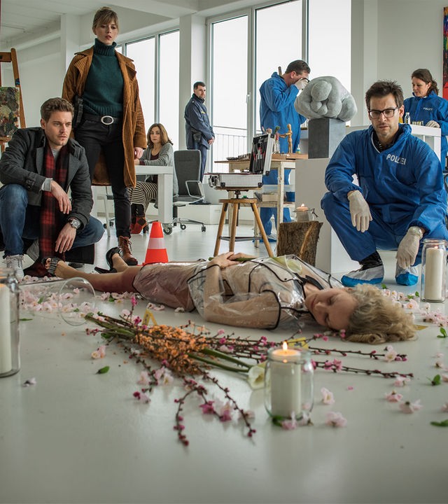 "SOKO München - Ein kunstvoller Tod": In einer Galerie liegt eine weibliche Leiche auf dem Boden um sie herum sind Blumen und Kerzengläser verteilt. Dr. Weissenböck (Florian Odendahl) sitzt in der Hocke neben ihr und schaut sie an. Am Fußende sitzt Dominik Morgenstern (Joscha Kiefer) in der Hocke, neben ihm steht Resi Schwaiger (Mersiha Husagic), sie schauen ebenfalls auf die Leiche. Im Hintergrund ist die Spusi (Komparsen) zu sehen.