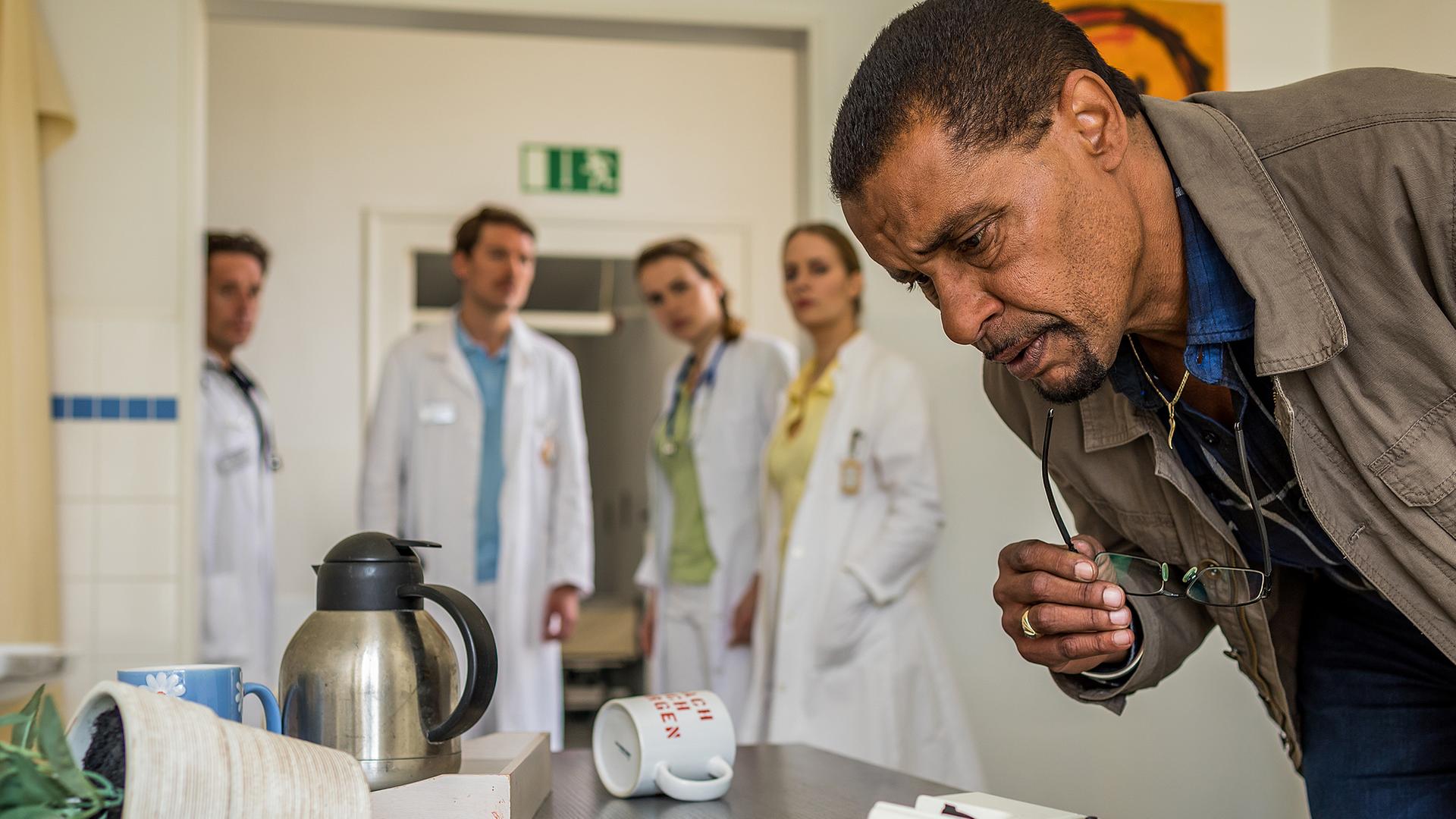 Franz Ainfachnur (Christofer v. Beau) steht vor einem Schreibtisch und schaut näher darauf