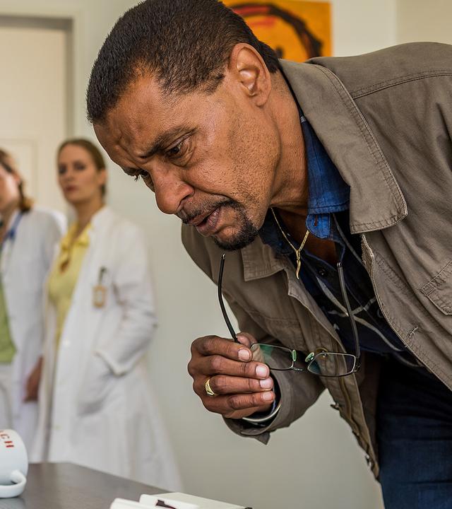 Franz Ainfachnur (Christofer v. Beau) steht vor einem Schreibtisch und schaut näher darauf