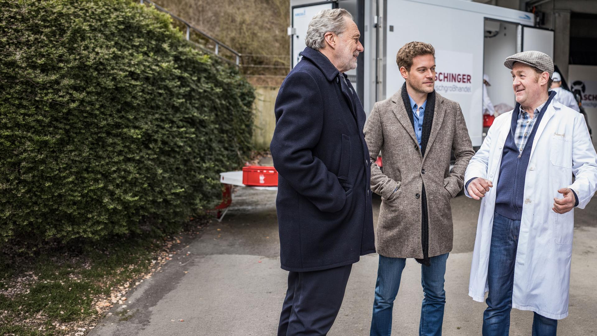 "SOKO München - Tödliche Vergangenheit": Die zwei Kommissare Arthur Bauer (Gerd Silberbauer) und Dominik Morgenstern (Joscha Kiefer) stehen mit einem der Giesinger Eiszapfen, Josef Aschinger (Jürgen Tonkel), vor seiner Metzgerei. Alle drei haben ein Lächeln im Gesicht. Die Kommissare haben die Hände in den Jackentaschen, während Josef Aschinger gestikuliert. Im Hintergrund ist ein Lieferwagen der Metzgerei zu sehen.