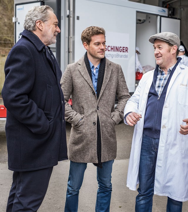 "SOKO München - Tödliche Vergangenheit": Die zwei Kommissare Arthur Bauer (Gerd Silberbauer) und Dominik Morgenstern (Joscha Kiefer) stehen mit einem der Giesinger Eiszapfen, Josef Aschinger (Jürgen Tonkel), vor seiner Metzgerei. Alle drei haben ein Lächeln im Gesicht. Die Kommissare haben die Hände in den Jackentaschen, während Josef Aschinger gestikuliert. Im Hintergrund ist ein Lieferwagen der Metzgerei zu sehen.