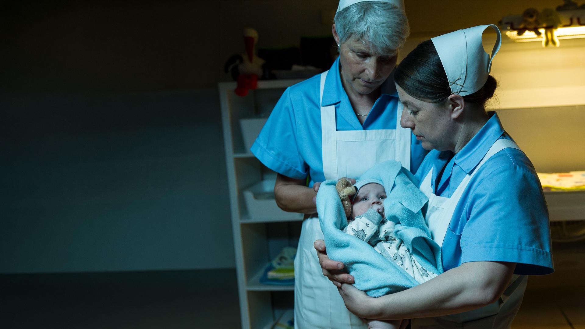 "SOKO Stuttgart - Babyklappe": Schwester Evi Neumann (Tilla Kratochwil) hält ein Neugeborenes (Komparse) in den Armen, während eine weitere Schwester (Komparsin) ihr assistiert. Beide tragen hellblaue Schwesterntracht mit weißen Schürze und weißen Häubchen. Sie stehen vor der geöffneten Babyklappe.