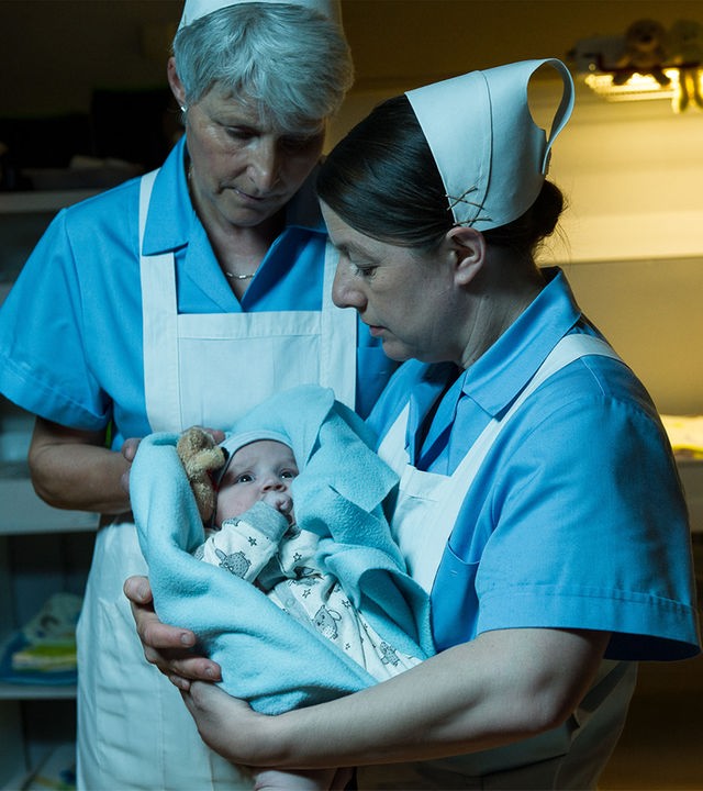 "SOKO Stuttgart - Babyklappe": Schwester Evi Neumann (Tilla Kratochwil) hält ein Neugeborenes (Komparse) in den Armen, während eine weitere Schwester (Komparsin) ihr assistiert. Beide tragen hellblaue Schwesterntracht mit weißen Schürze und weißen Häubchen. Sie stehen vor der geöffneten Babyklappe.