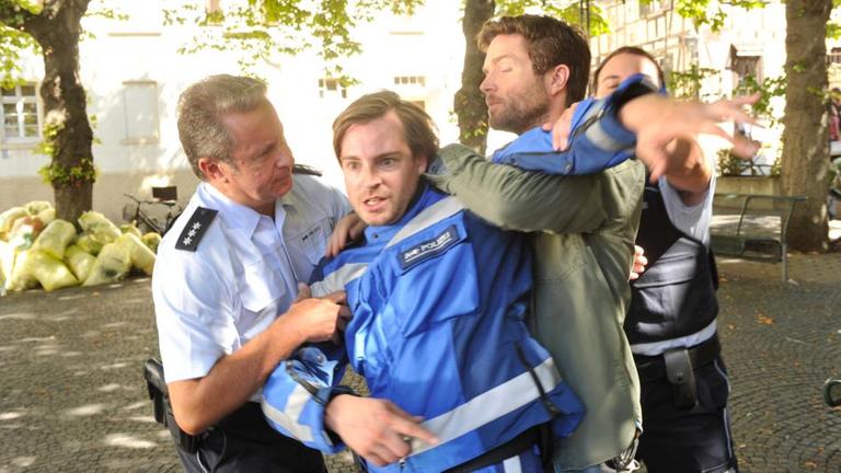 "SOKO Stuttgart - Major Mimis Ende": Jo Stoll (Peter Ketnath) und zwei Kollegen (Komparsen) überwältigen einen weiteren, ziemlich wütenden Kollegen auf einem von Bäumen umgebenen Platz. Es handelt sich um Ronny Schröder (Robin Czerny).