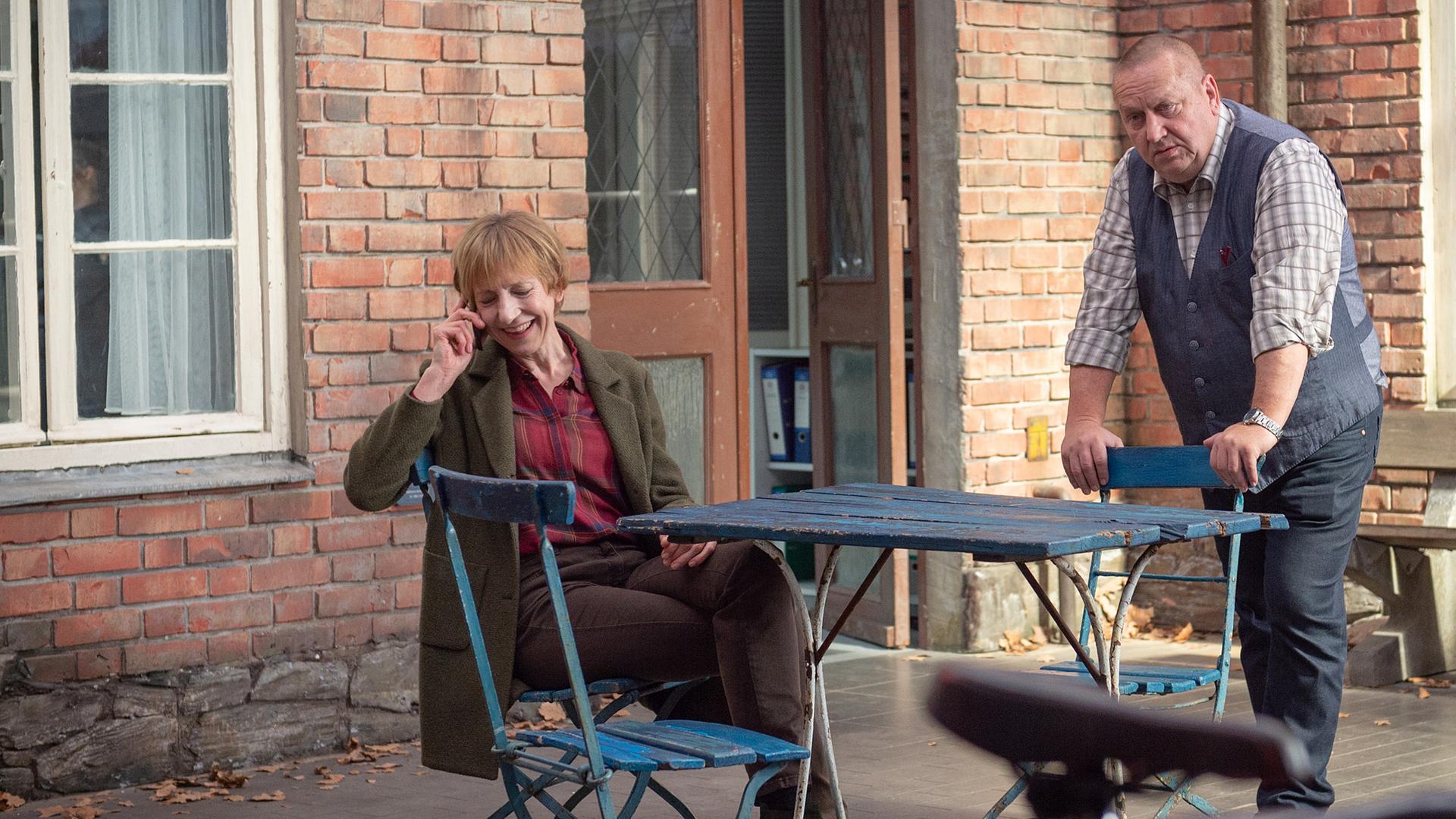 Helene (Katharina Blaschke) sitzt telefonierend an einem Tisch im Hof des Reviers. Bei ihr ist Reuter (Udo Kroschwald), der sich auf einer Stuhllehne abstützt
