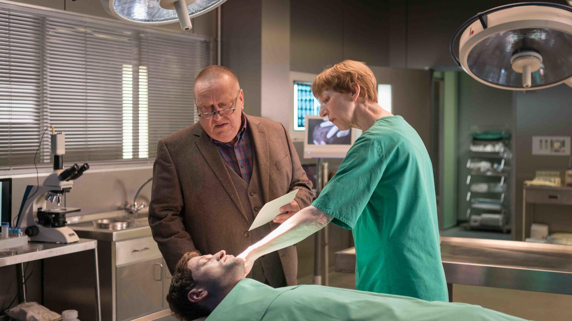 Kriminalkommissar Jan Hinrich-Reuter steht in einem braunen Anzug neben Dr. Helene Sturbeck, die einen Laborkittel trägt, in einem Obduktionssaal. Sie stehen vor einem männlichen Leichnam und Dr. Sturbeck zeigt auf eine Stelle am Kopf des toten Mannes.