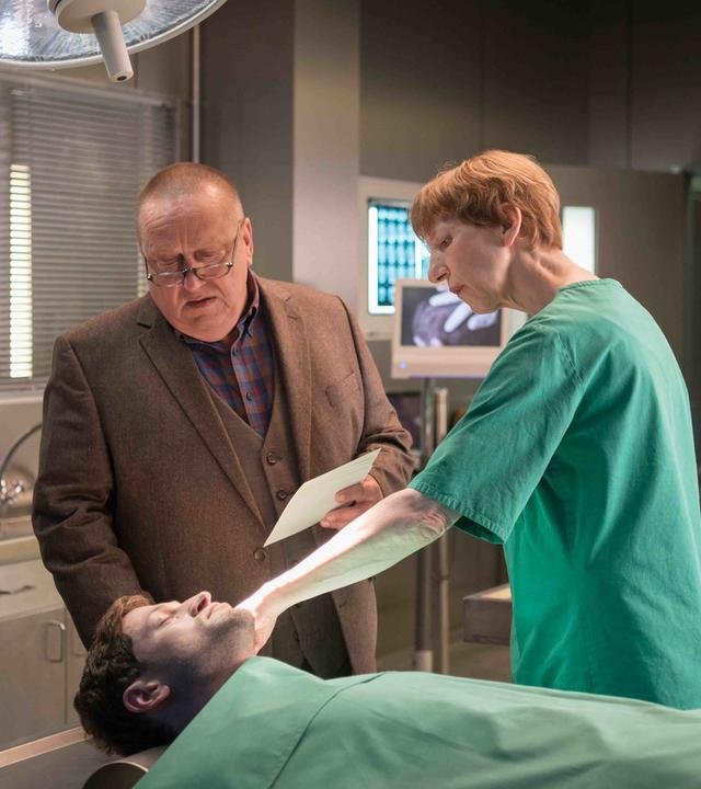 Kriminalkommissar Jan Hinrich-Reuter steht in einem braunen Anzug neben Dr. Helene Sturbeck, die einen Laborkittel trägt, in einem Obduktionssaal. Sie stehen vor einem männlichen Leichnam und Dr. Sturbeck zeigt auf eine Stelle am Kopf des toten Mannes.