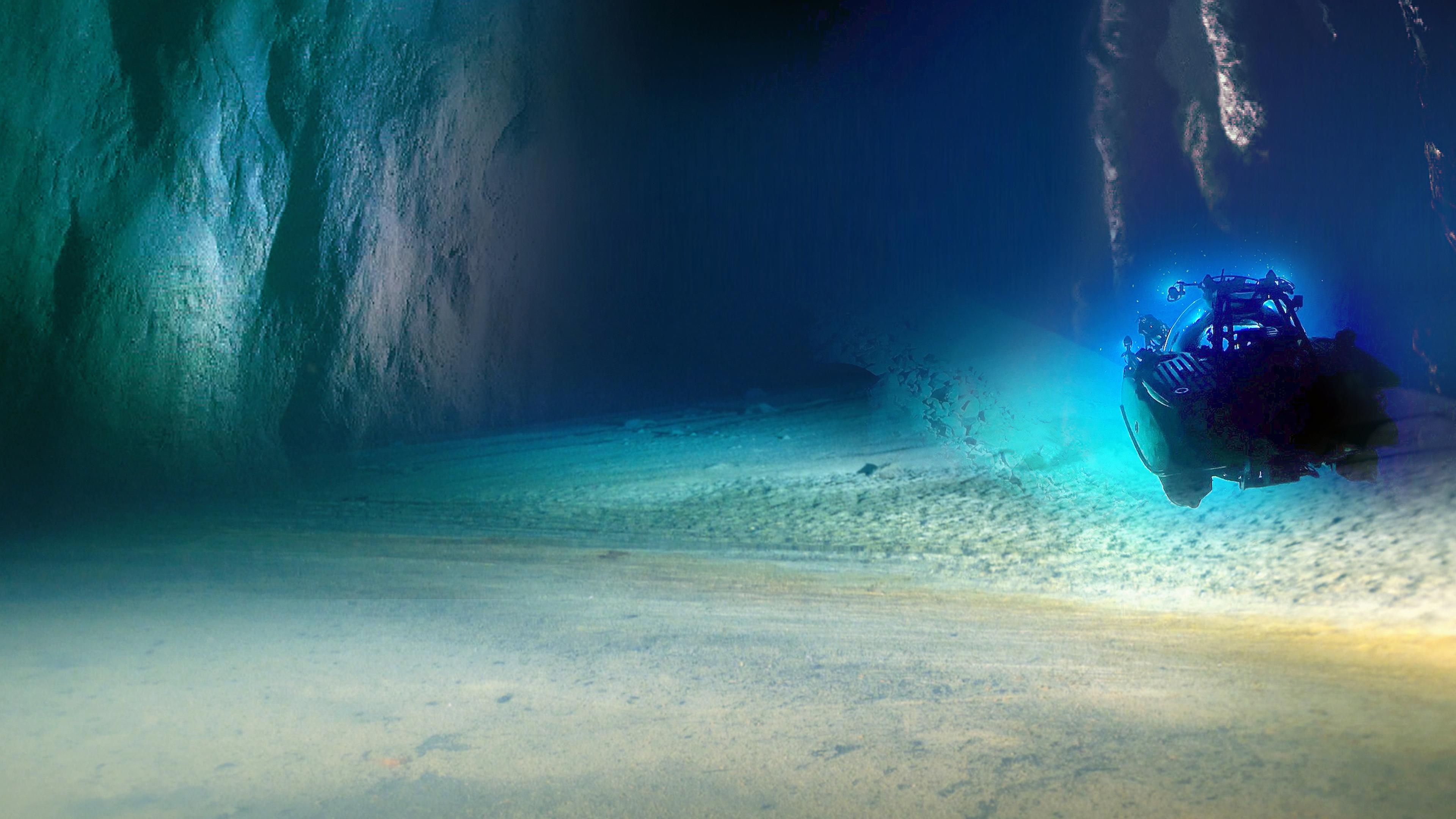 Blick auf türkis und blau leuchtende Felswände und einen Strand unter Wasser. Ein Tauchboot untersucht den Meeresgrund.