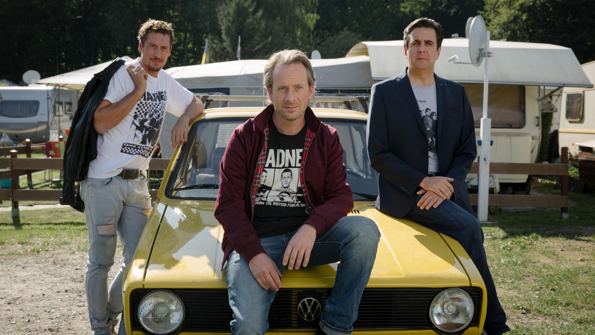 "Der Sommer nach dem Abitur": Paul (Hans Löw), Ole (Fabian Busch) und Alexander (Bastian Pastewka). Ole und Alexander sitzen entspannt auf der Motorhaube eines alten gelben Golfs während Paul links an den Wagen gelehnt steht.