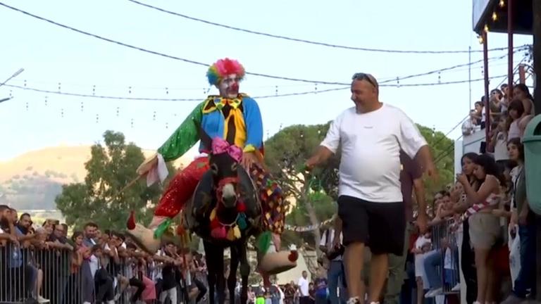 Ein Esel wird von einem Mann geritten, der als Clown verkleidet ist. Der Esel ist ebenfalls bunt geschmückt.