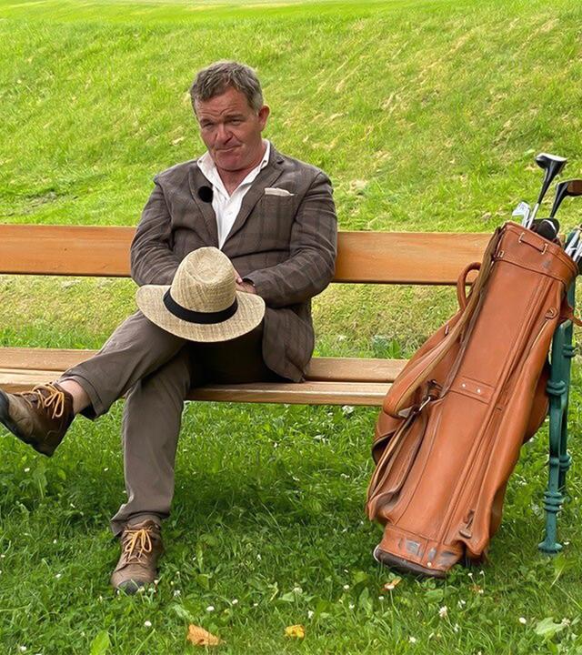 Das Bild zeigt einen Mann, der auf einer Bank sitzt. Er trägt ein kariertes Jackett, ein Hemd und eine Hose. Auf seinen Knien hält er einen Strohhut, der mit einem schwarzen Band verziert ist. Neben ihm steht eine Golf-Tasche aus Leder, aus der mehrere Golfschläger herausragen. Im Hintergrund ist eine grüne Rasenfläche zu sehen, die sanfte Hügel aufweist. Das gesamte Szenario vermittelt eine entspannte, sommerliche Atmosphäre.